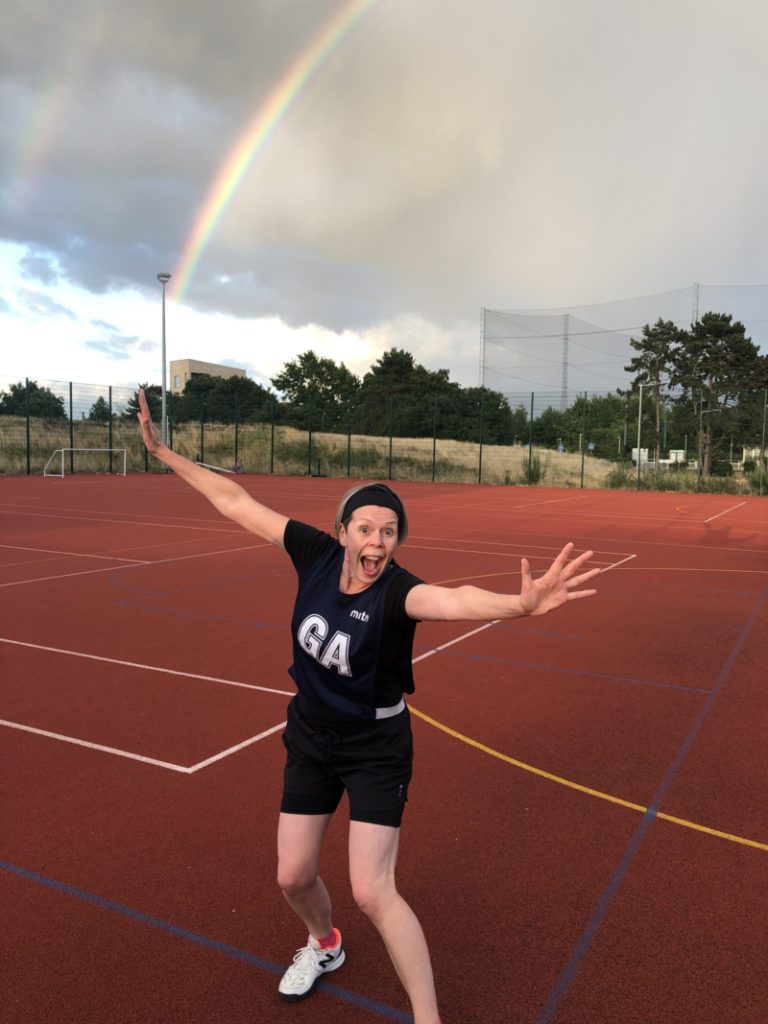Woman jumping and smiling in her netball kit with a rainbow in the sky