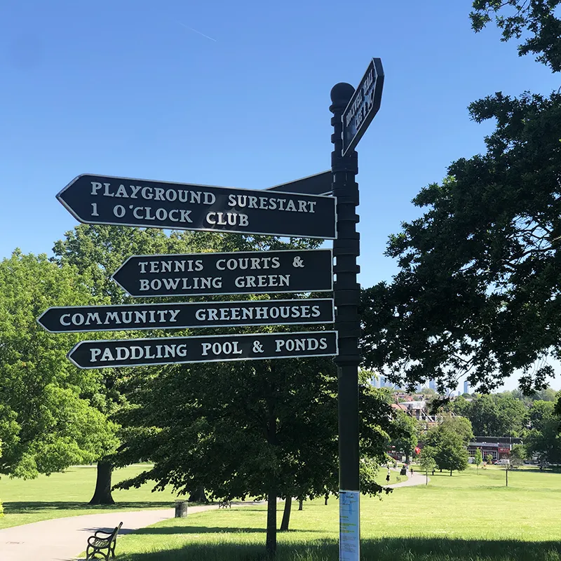 Park signpost showing directions to playground, tennis courts, greenhouses, and paddling pool on a sunny day.